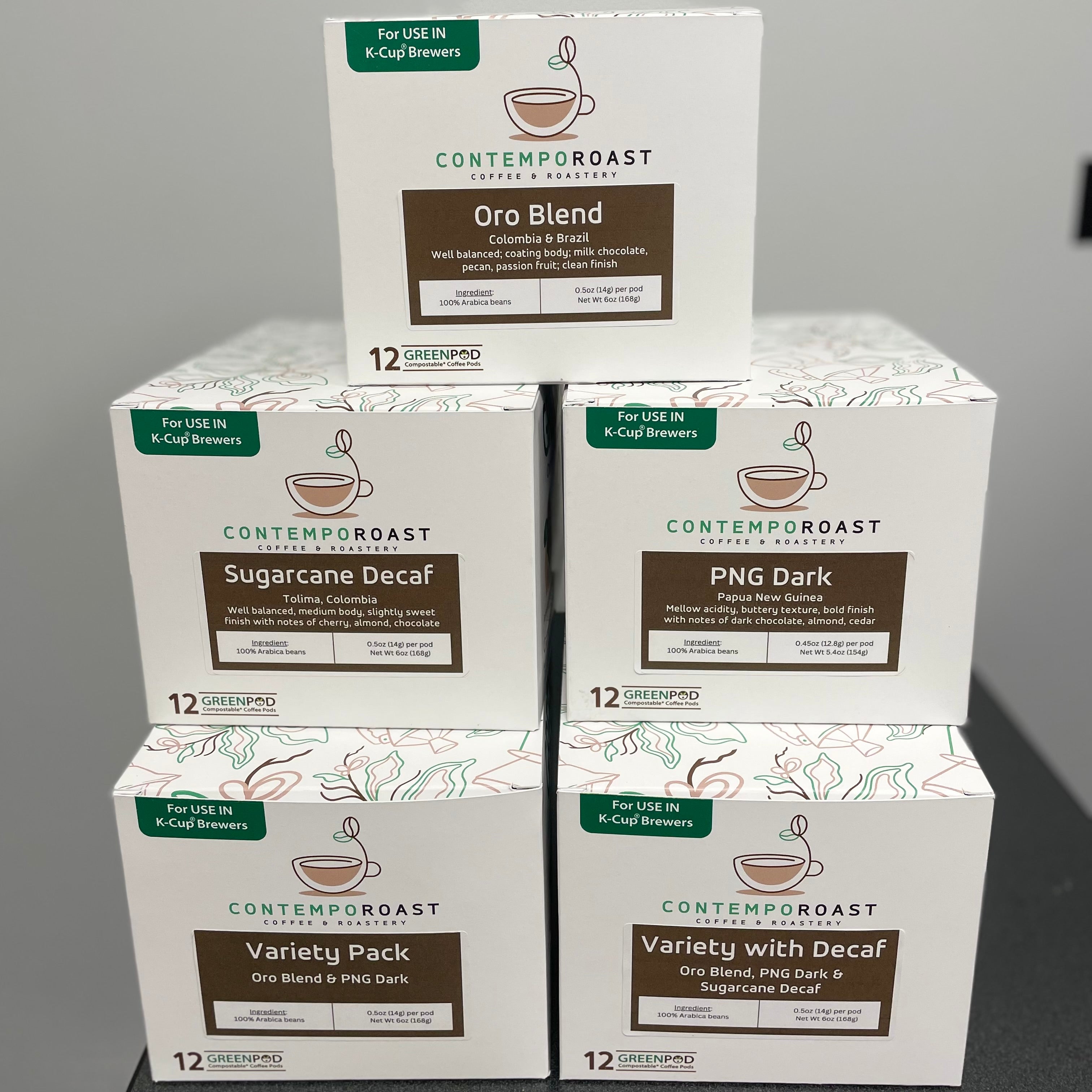 A stack of fully compostable coffee pod boxes, showing the five different options: Oro Blend; Sugarcane Decaf; PNG Dark; Variety Pack (Oro Blend & PNG Dark); and Variety with Decaf (Oro Blend, PNG Dark, Sugarcane Decaf)