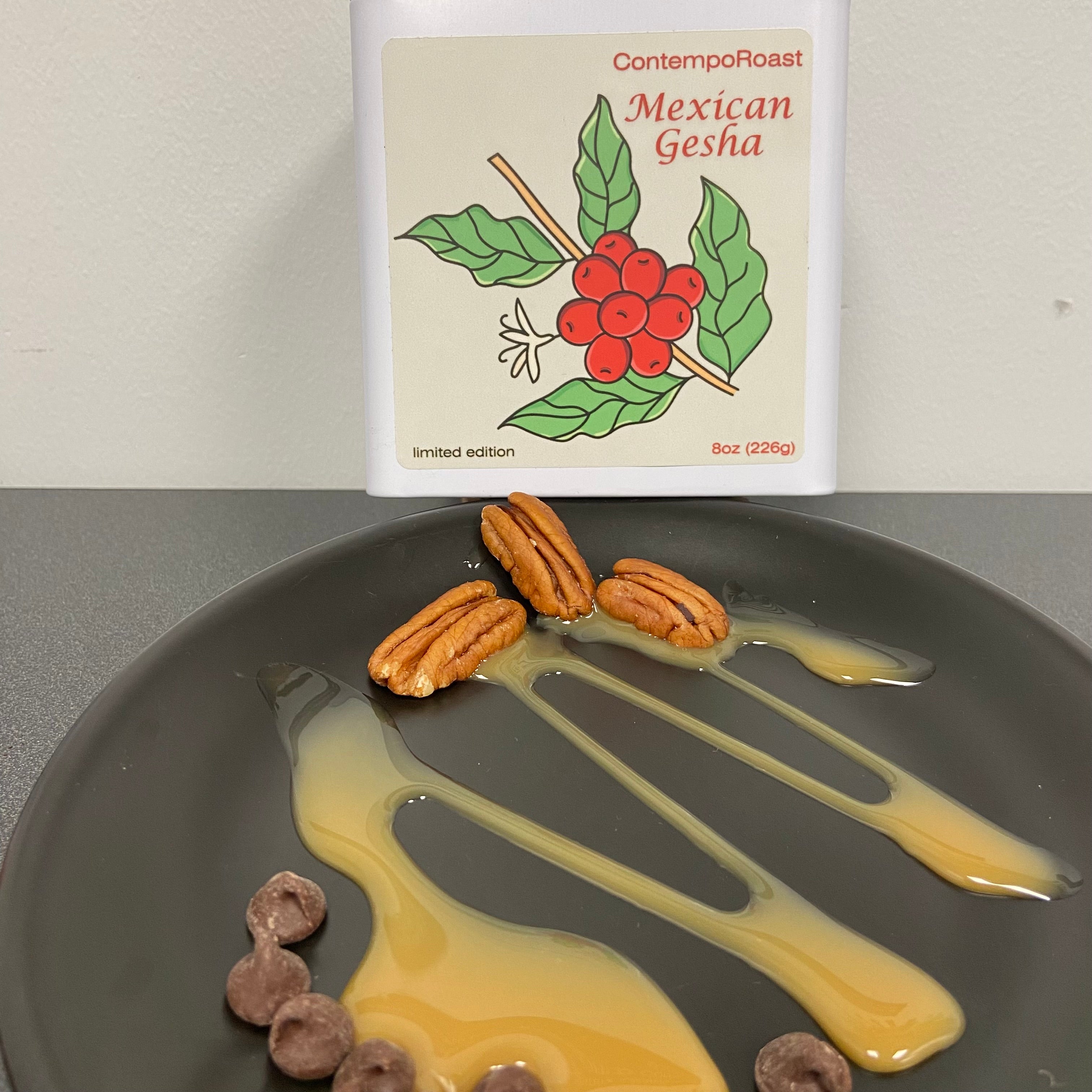 A plate with pecans, chocolate chips and caramel sauce in the foreground with a keepsake tin of Mexican Gesha coffee in the background