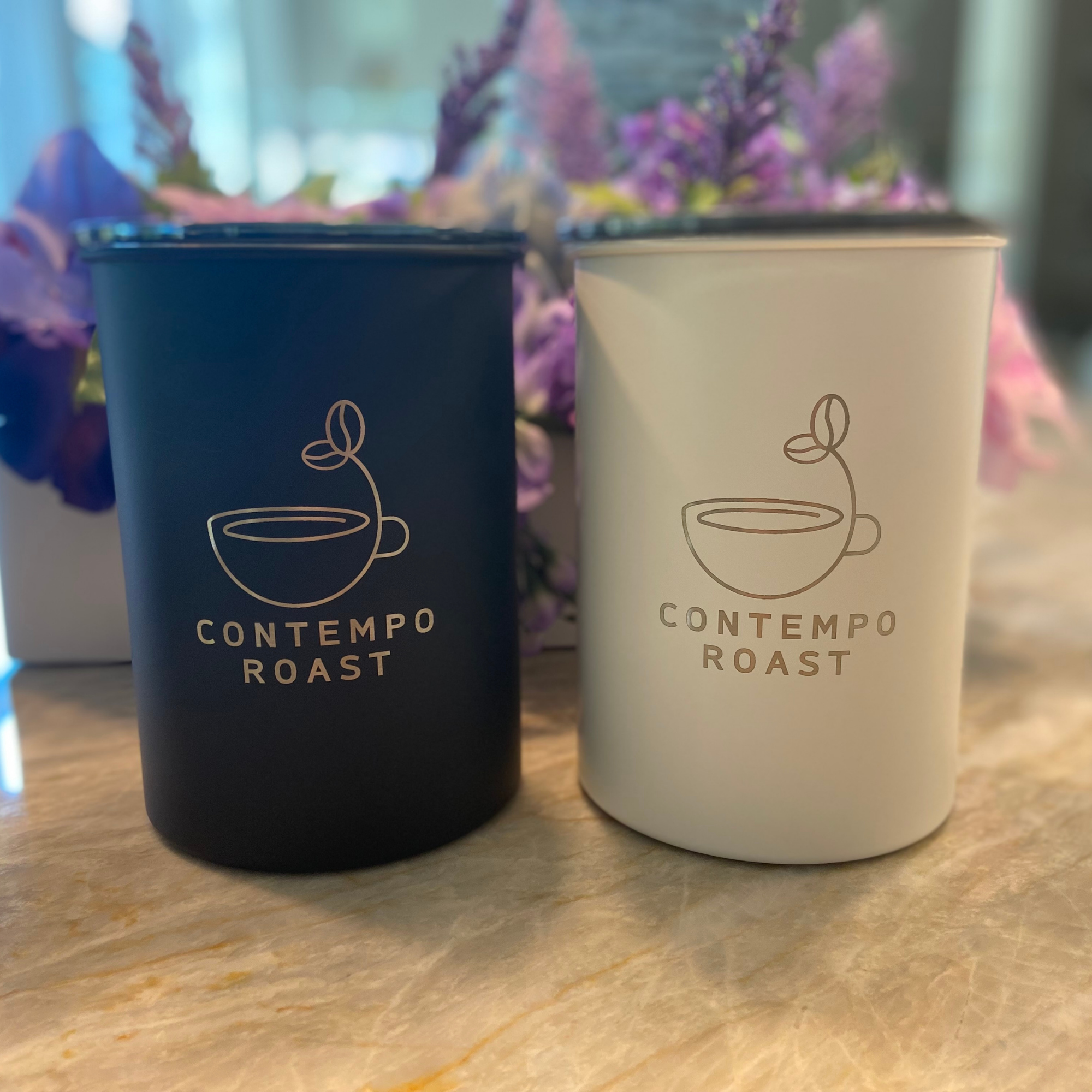 matte black and matte white canisters, both etched with the ContempoRoast logo, sitting side-by-side on a counter in front of purple flowers